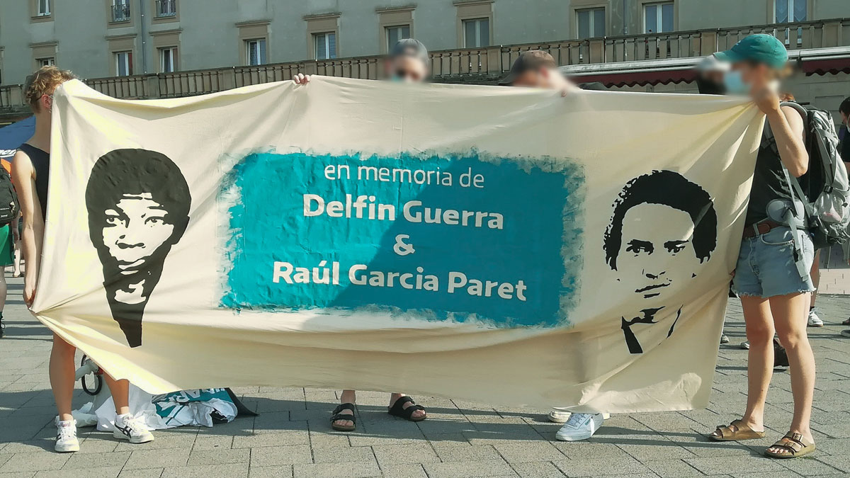 Auf dem bahnhofsvorplatz in Merseburg halten Menschen ein Banner hoch. Darauf steht: "en memoria de Delfin Guerra & Raúl Garcia Paret". Rechts und links von der Schrift sind die Gesichter von Delfin und Raúl abgebildet.
